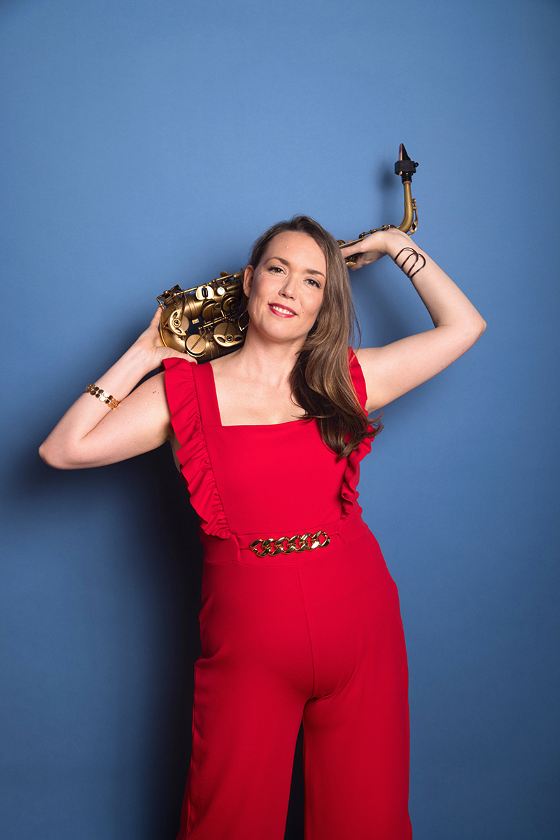 Erica von Kleist wearing an orange jumpsuit holds her sax behind her head